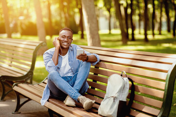 Fototapeta premium Funky black guy sitting on bench and listening to music on cellphone at city park