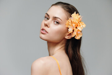 Fototapeta premium Young woman with long hair and orange flower in her hair, showcasing natural beauty and elegance with soft expressions on a neutral background.