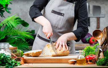 Chef cutting white bread. Cozy kitchen with wooden table, kitchenware and ingredients for cooking. Homemade baking, cooking, food blogging