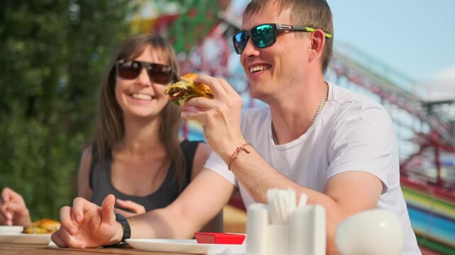 A couple sits at a summer restaurant table, smiling and enjoying their weekend at an amusement park. The man eagerly eats a burger while the woman, wearing sunglasses, keeps the conversation flowing.