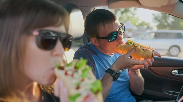 Side view, road trip date. The woman in sunglasses eats a hot dog with lettuce and cheese sauce, while the man holds his snack with both hands. The sunset glow highlights the romance of their journey.
