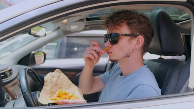 Side view through the car window. A man in a blue t shirt and sunglasses sits in his parked car, holding a paper of fries, sniffing one before popping it into his mouth and savoring the taste.