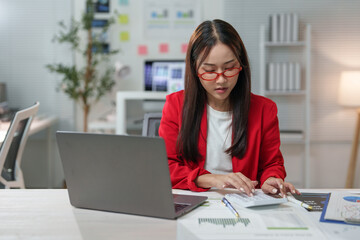 Fototapeta premium Focused businesswoman in a modern office setting, using a calculator and reviewing financial documents. A laptop and charts are visible, indicating a detailed analysis of data