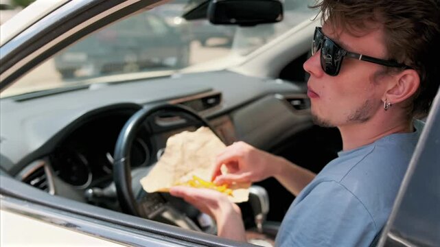 Side view through the car window. A man in a blue T shirt and sunglasses sits behind the wheel, holding a packet of fries and biting into one, enjoying his snack and the warm summer day outside. 