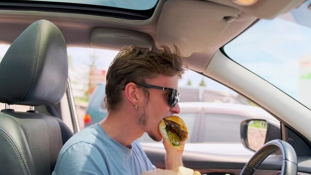 Low angle A young man in sunglasses sits in his car, biting into a burger with pickles, vegetables, and cheese sauce. He holds it over a wrapper as a slice of tomato slips out and falls to the floor.