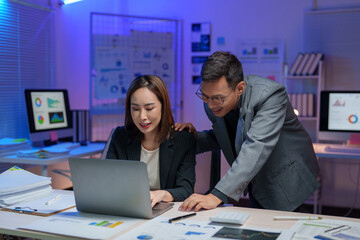 Two business professionals collaborate in a modern office, analyzing charts and graphs on a laptop. The workspace is illuminated with blue lighting, enhancing the tech-savvy atmosphere