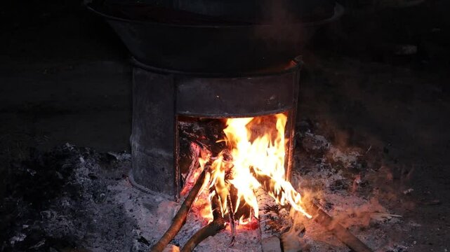 Traditional charcoal stove burning. Bright orange-yellow flames are emitted from the firebox in a cylindrical metal stove. The stove is set on the ground with scattered ashes and wood chips.