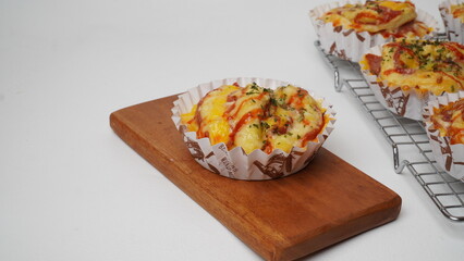 A group of salty bread is served in a paper cup with smoked beef filling, orange cheddar and topped with mayonnaise, tomato sauce, and parsley which is placed on a cooling rack with a white background