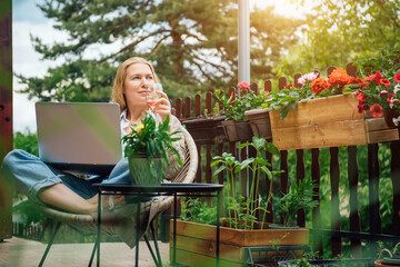 Young adult Caucasian blonde woman sitting on cottage terrace with a glass of rose wine and using laptop, online work in the countryside concept.  Female freelancer does business in Her home