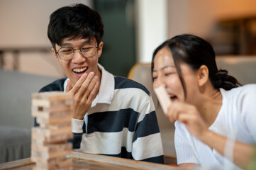 Two cheerful Asian friends, a male and a female, enjoy playing Jenga together at home.