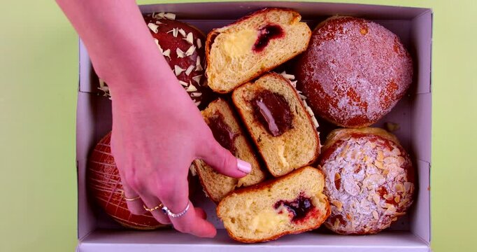 A hand selecting from a box of assorted Berliner doughnuts, perfect for Carneval, featuring chocolate, custard, and jam fillings with sugar, glaze, and almond toppings. Donut on green background.
