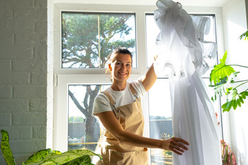 Woman in apron hangs transparent tulle curtains on large windows in the house inside the interior...