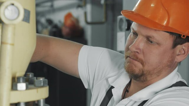 A worker in a safety helmet at a factory tightens a nut on a pipe with a large wrench. The wrench tightens the nut. A worker repairs a pipeline.
