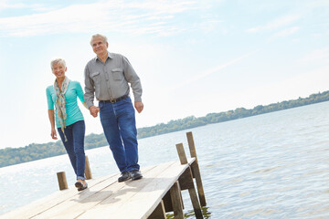Fototapeta premium Happy, elderly and couple walking on pier for retirement, vacation journey and travel destination. Below, senior people and holding hands for marriage commitment, holiday location and bonding on lake