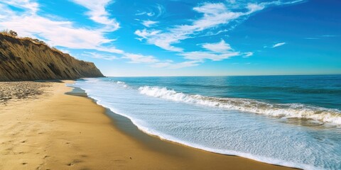 Fototapeta premium Serene beach landscape with golden sand and gentle waves, clear blue ocean under a vibrant sky with wispy clouds, cliffs in the background.