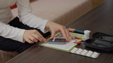A woman sits at a table, preparing to test her blood sugar level. She inserts a test strip into a glucometer while organizing her diabetes monitoring supplies.
