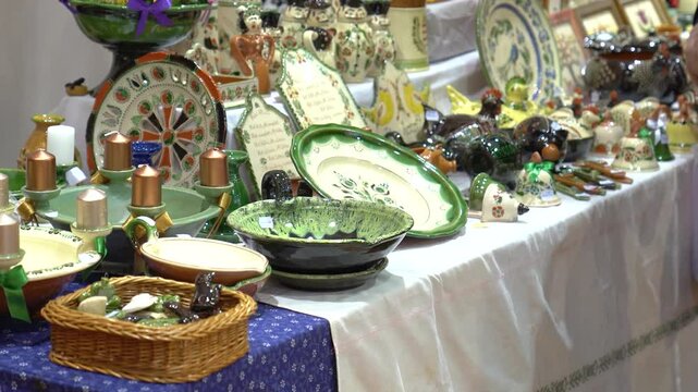A left-to-right pan shot in a thrift store displaying handcrafted porcelain plates and figures. A person examines a small porcelain pig on a table with patterned tablecloths.