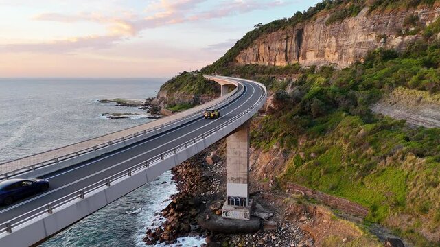 Aerial tracking footage of a car along the Sea Cliff Bridge, highlighting the breathtaking road suspended over the sea. Cinematic drone footage.