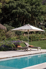 white sunshade casts a shadow over two cushioned lounge chairs near swimming pool. In the background, a lush, green garden, featuring various plant species Tranquility and relaxation.