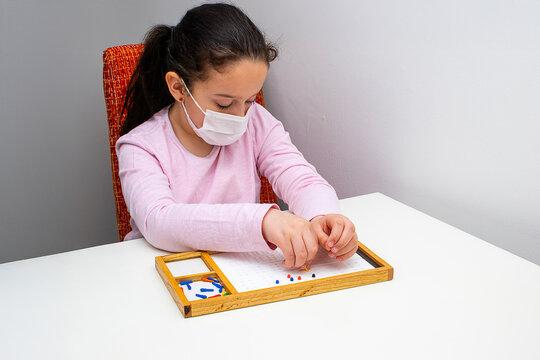 A blind girl wearing a protective mask practices reading and writing with a Braille learning board at home. Her hands carefully navigate the tactile symbols, demonstrating the significance of touch in