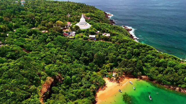 Drone pullback to reveal the Japanese Peace Pagoda surrounded by greenery and the ocean in Sri Lanka with tourists at beach below