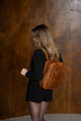 A girl in a warm sweater, a black short skirt and nylon tights is posing with a brown backpack.