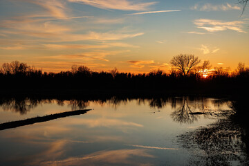 panoramica su un ambiente naturale fluviale al tramonto, con la luce solare che colora di arancione il cielo azzurro, leggermente nuvoloso, e si specchia sull'acqua ferma