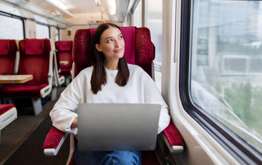 Dreamy lady freelancer sitting in train and looking out of window, enjoying the view while using...