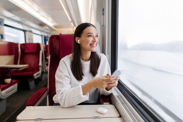 Happy woman sitting on seat in train, listening to music or audiobook in earphones and using smartphone, lady enjoying travel