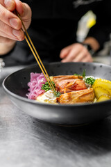 A close-up of a chef using chopsticks to garnish a gourmet dish in a stylish black bowl. The plate features grilled chicken, pickled onions, and fresh herbs on a rustic metal surface.