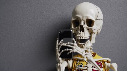 Skeleton posing with smartphone, taking playful selfie against neutral gray background, representing modern digital culture with humorous dark twist