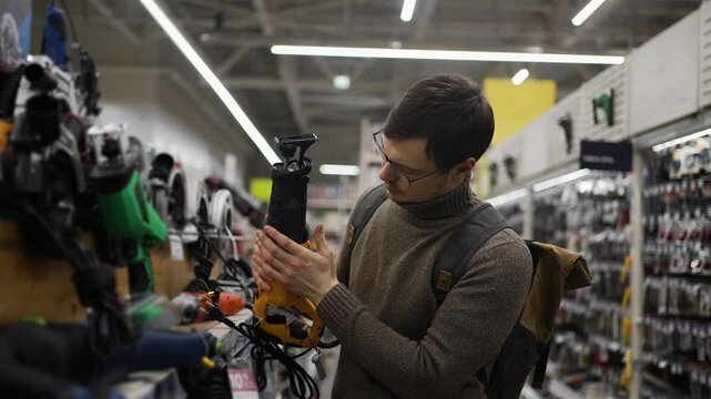 Young male customer comparing reciprocating saws, carefully examining electric power tools while browsing shelves in well-lit hardware store retail space, wearing backpack and glasses