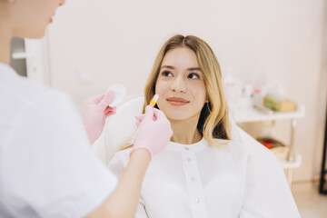 Fototapeta premium Cosmetologist wearing pink gloves holding syringe and cotton pad, preparing botox injection for smiling woman in white shirt