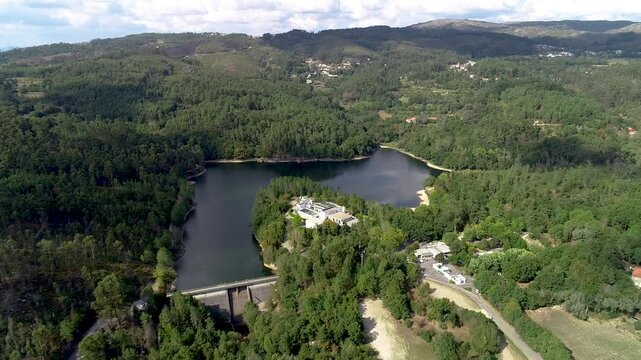Distant drone shot of a dam and the respective river from both sides. There is also a building by riverside, and many kilometers of forest, that extends into every direction.