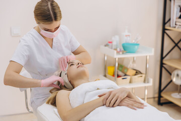 Fototapeta premium Beautician wearing surgical mask and gloves injecting botox on forehead of young woman for facial rejuvenation treatment in beauty salon