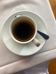 Top view of a white cup filled with black coffee on a saucer with a spoon, placed on a white tablecloth.