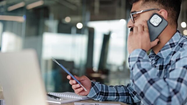 Young businessman confirming travel booking via phone call. Handsome male making business trip arrangements using passport and ticket in modern office.