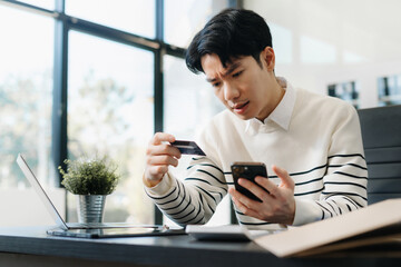 Young Asian man stressed over credit card debt, holding bills in frustration. Financial anxiety and...