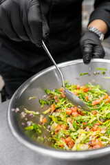 A close-up of a person in black gloves mixing a fresh salad in a stainless steel bowl, featuring...