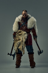 Full-length portrait of man as medieval warrior, Viking in full armor and fur elements, standing with two axes in fierce pose. Battlefield, determination. Concept of historical portrait, courage