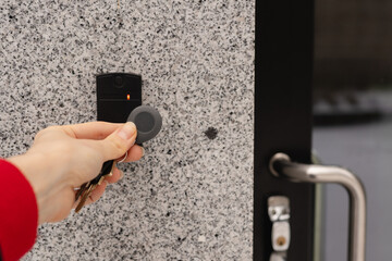 Close-up of a female hand holding a keychain with a key fob near a key reader access device on a building’s exterior wall with door. modern security, smart access, and contactless entry technology