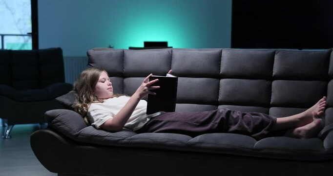 Young girl lies on sofa reading interesting book in modern living room. Schoolgirl enjoys hobby discovering enjoyable novel in evening slow motion