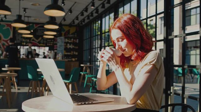 Young woman with red hair is working on her laptop while sitting in cafe drinking coffee. Close-up. Comfortable freelancing, good enough.
