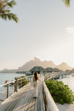 A woman in a flowing white dress strolls along a wooden boardwalk towards overwater bungalows in Bora Bora, bathed in golden sunset light. A serene moment in a paradise of turquoise waters