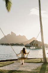 A woman in a flowing white dress swings under a palm tree at sunset, overlooking the turquoise...