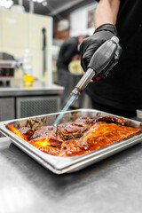 A chef with black gloves uses a culinary torch to caramelize sauce on a tray of grilled meat in a professional kitchen setting. The focus is on the cooking technique and delicious food.