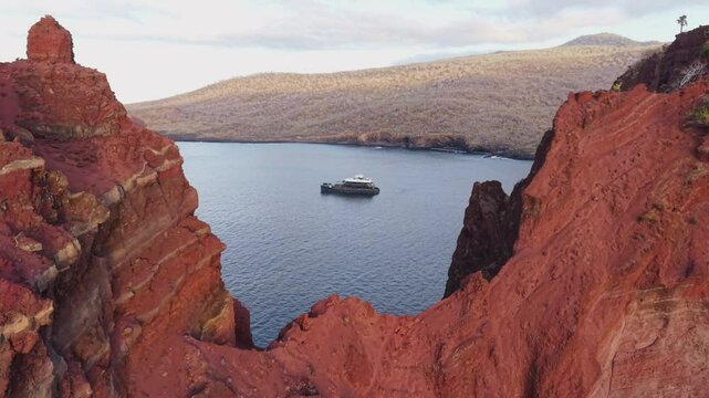 Towering red volcanic cliffs overlooking tourist yacht gliding through crystal clear Galapagos waters, showcasing deep azure ocean backdrop