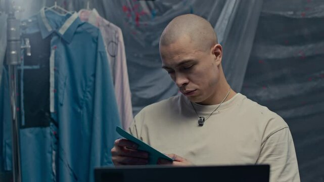 Medium close up of young Caucasian man sitting at table in front of laptop in painting studio covered with transparent sheeting choosing patterns to redesign shirts hanging on stand in background