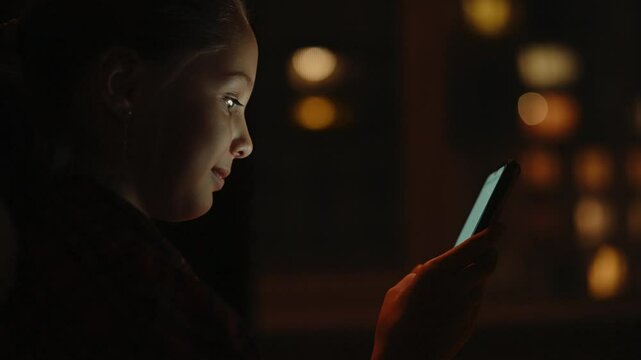 Close-up face of cute young 10 yo girl using smartphone at night sitting alone in dark bedroom, face illuminated by screen, city lights reflecting in window, creating cozy and intriguing atmosphere.