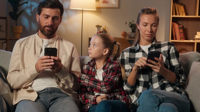Caucasian family parents mother father using mobile phone smartphones scrolling gadgets addict dependence internet ignore daughter child sad kid girl ask attention lonely problem at evening home night